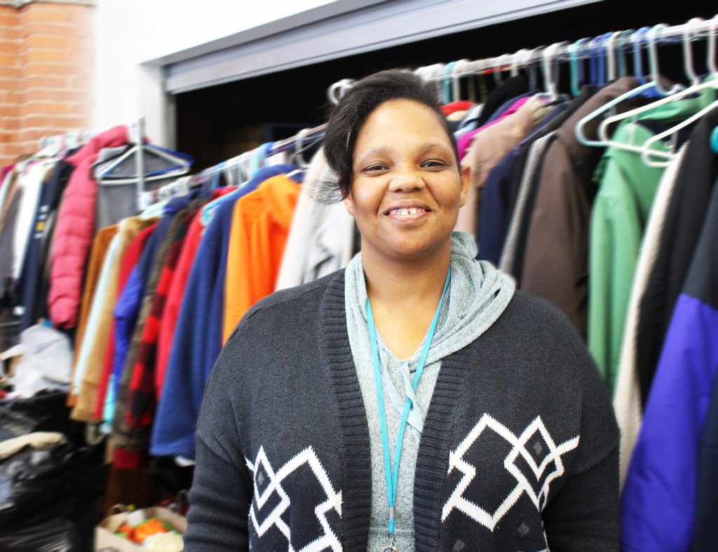 La Toyei smiles while standing in front of multiple jackets hanging on racks at city rescue mission