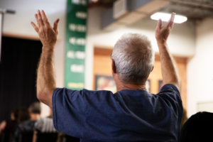 A elderly man in a blue shirt holds his hands up in praise