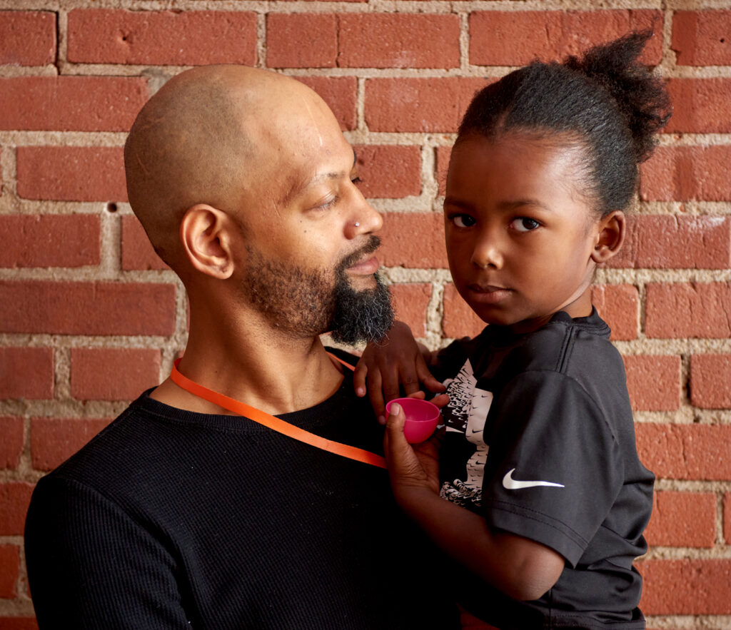 A man and his young daughter in front of a red brick back drop at city rescue mission