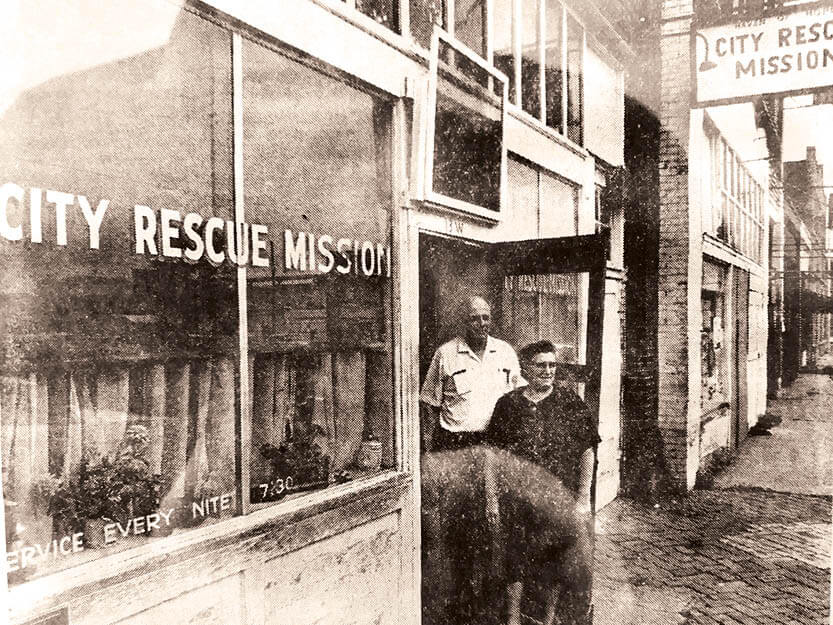 An old black and white historical photo of two members of city rescue mission standing outside the original building