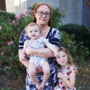 A young mother with red hair holds a young infant and stands by her younger daughter